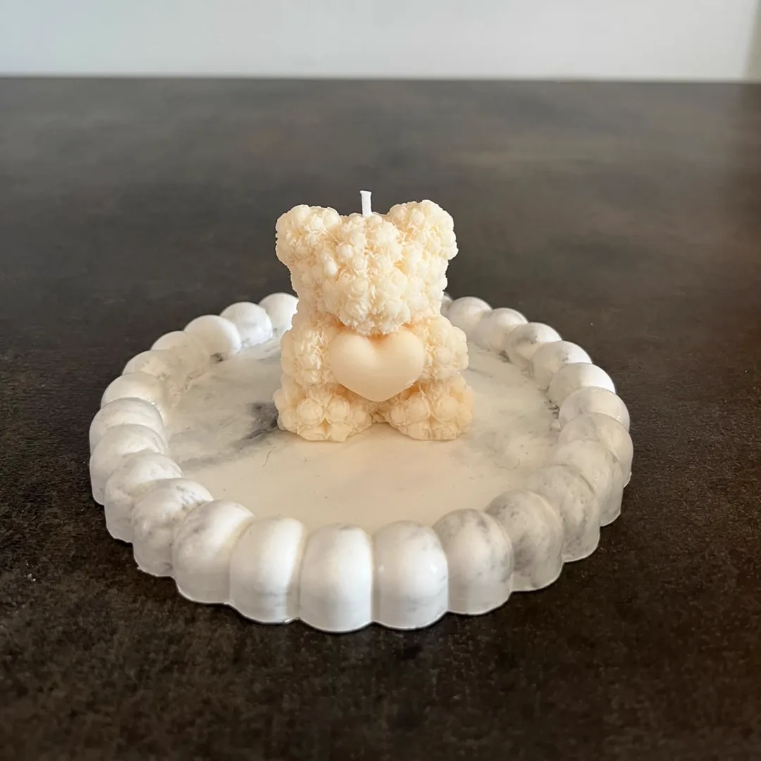 Vela de soja con forma de osito texturado abrazando un corazón, sobre bandeja de yeso